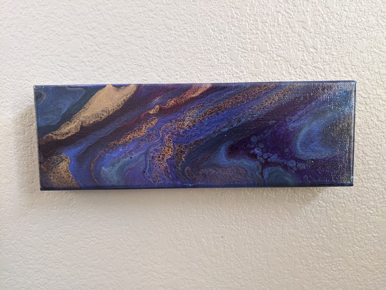 Puede incluir: Pintura abstracta sobre lienzo con colores azules, morados y dorados en espiral. La pintura tiene un aspecto texturizado y fluido.