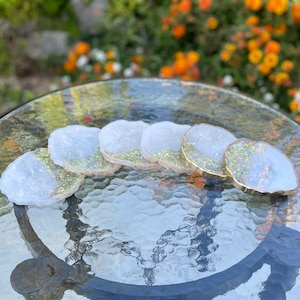 May include: Set of six white resin coasters with gold glitter and a gold rim. The coasters are shaped like irregular circles and are arranged in a semi-circle on a glass table.