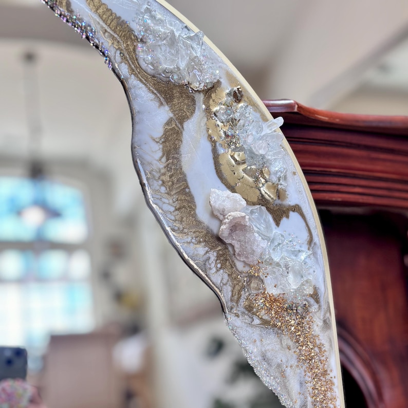 May include: A decorative mirror frame featuring a curved edge adorned with white and gold resin swirls, embedded with clear and pale pink crystalline mineral clusters, and accented with fine gold glitter particles along the textured surface.
