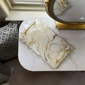 May include: A decorative rectangular tray with a white and gold marbled design. The tray has gold handles and is embellished with clear beads. The tray is on a white surface.