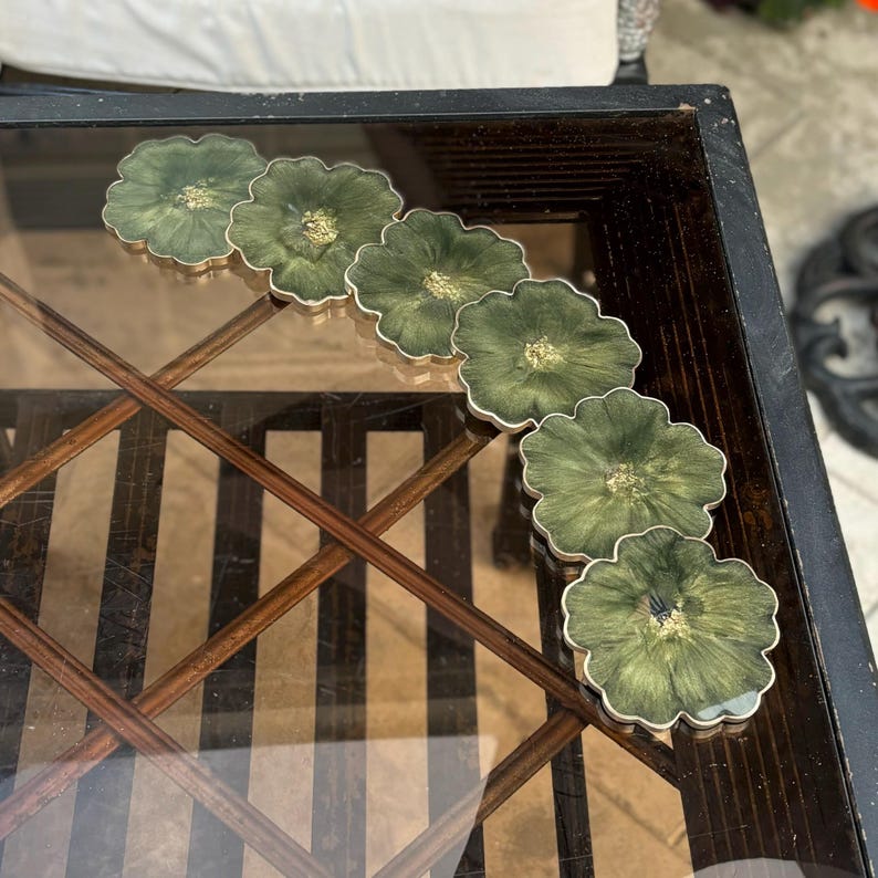 Peut inclure: Ensemble de six sous-verres en r&eacute;sine verte et dor&eacute;e avec un motif floral. Les sous-verres sont dispos&eacute;s en demi-cercle sur une table en verre avec un cadre en bois.