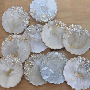 May include: A selection of handcrafted resin coasters in various shapes. The coasters feature a pearlescent white and gold marbled design, embellished with faux pearls, rhinestones, and floral accents. The coasters are arranged on a wooden surface.