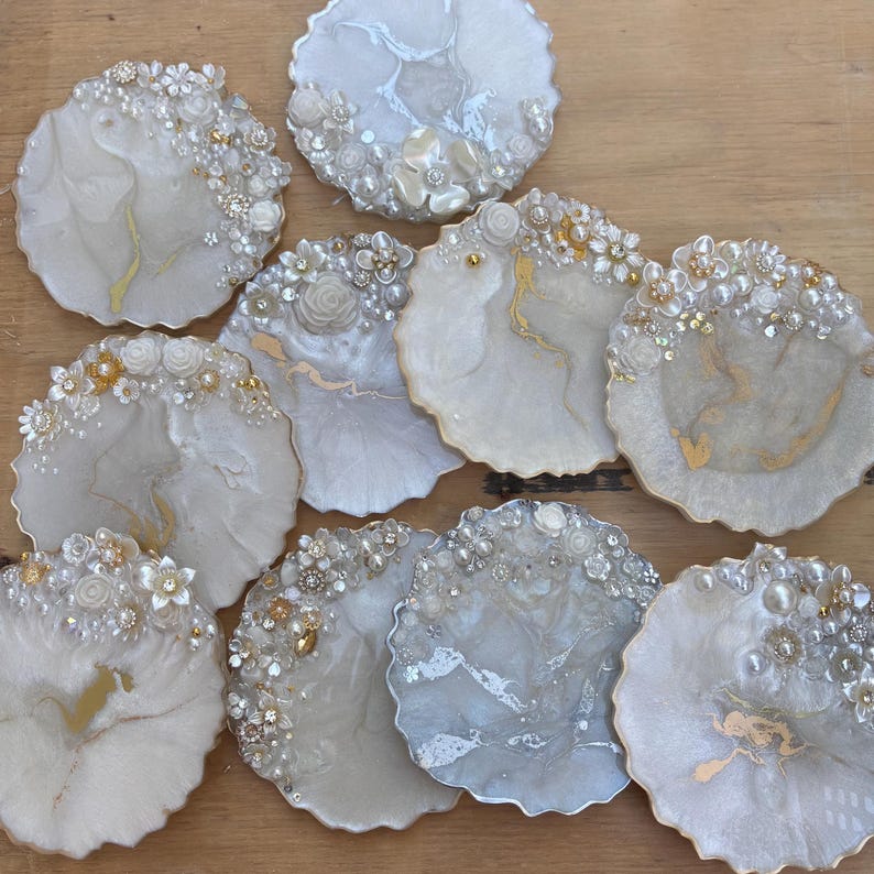 May include: A selection of handcrafted resin coasters in various shapes. The coasters feature a pearlescent white and gold marbled design, embellished with faux pearls, rhinestones, and floral accents. The coasters are arranged on a wooden surface.