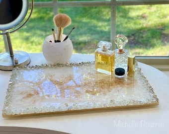 Puede incluir: Una bandeja de resina blanca con destellos dorados y un borde de cristal transparente. La bandeja está llena de pequeños cristales transparentes y tiene dos botellas de perfume de vidrio y un pequeño recipiente negro en la parte superior.