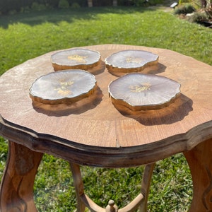 May include: Four white and gold resin coasters with a unique, irregular shape. The coasters are on a wooden table with a carved design.