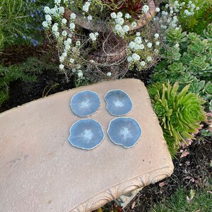 K&ouml;nnte beinhalten: Vier blaue und wei&szlig;e Untersetzer aus Kunstharz mit goldfarbenem Rand liegen auf einer Steinbank. Die Untersetzer haben eine einzigartige, organische Form. Der Hintergrund zeigt einen Garten mit verschiedenen Pflanzen und Blumen.