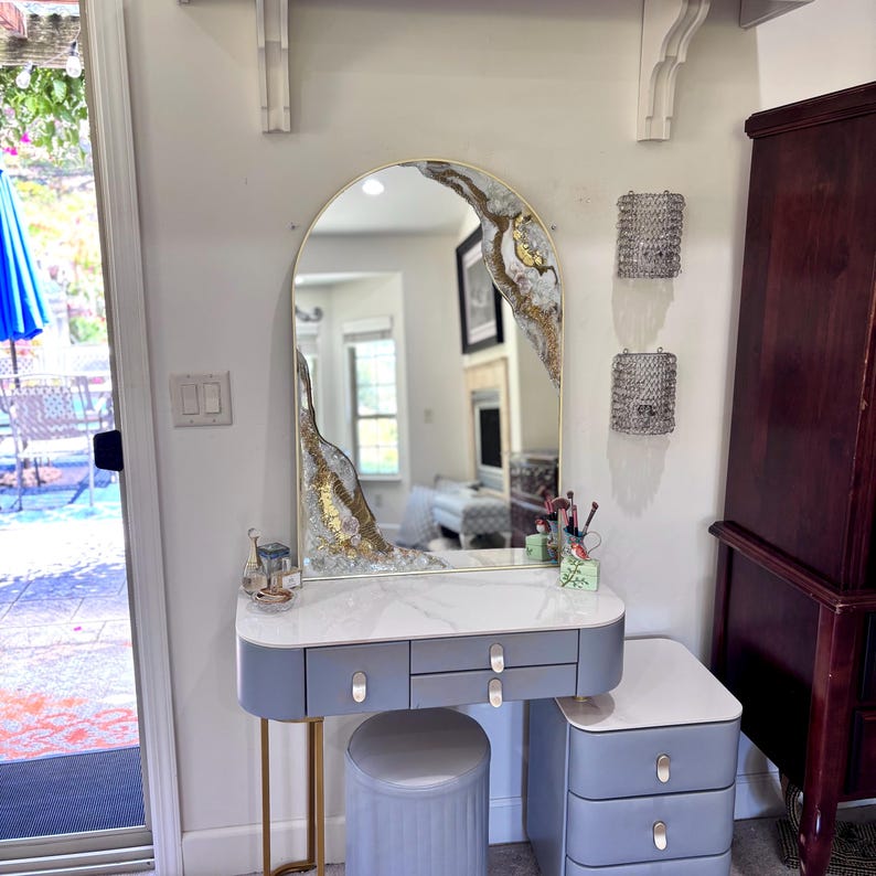 May include: A vanity set featuring a gray desk with three drawers, a matching three-drawer side cabinet, and a cylindrical stool. An arched mirror with gold trim and decorative white and gold resin accents sits atop the white marble-patterned desk.