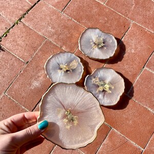 May include: Four pink and white resin coasters with gold leaf accents and a gold rim. The coasters are on a brick surface.