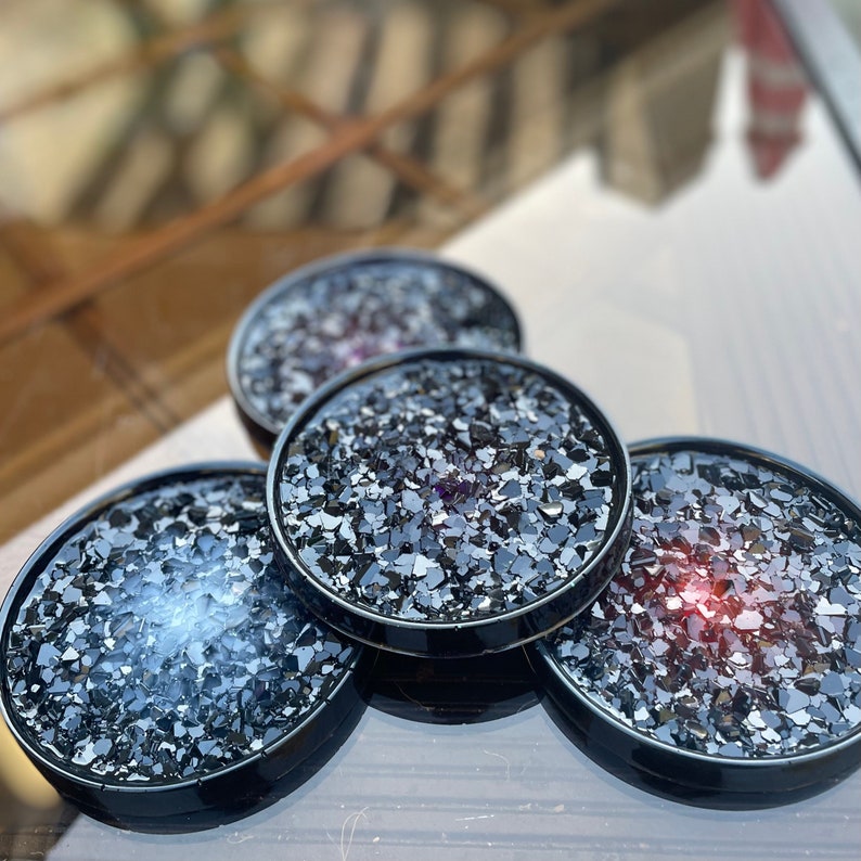 May include: Four black coasters with a glittery, iridescent finish. The coasters are round and have a raised edge. They are made of resin and have a unique, sparkly look.