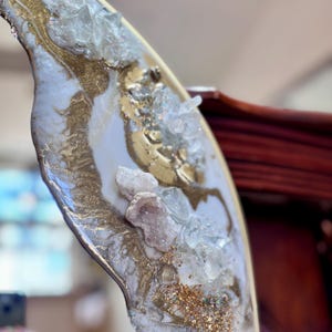 May include: A decorative resin art piece featuring a swirling white and gold marbled pattern. The surface is embellished with clusters of clear, jagged quartz crystals and fine gold glitter, creating a textured, multi-dimensional appearance.
