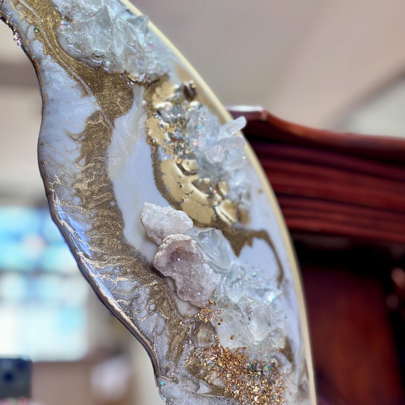 May include: A decorative resin art piece featuring a swirling white and gold marbled pattern. The surface is embellished with clusters of clear, jagged quartz crystals and fine gold glitter, creating a textured, multi-dimensional appearance.