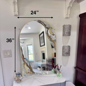 May include: An arched wall mirror with a gold frame, featuring decorative resin accents in white, gray, and gold leaf along the top right and bottom left corners. The mirror measures 24 inches in width and 36 inches in height as indicated.