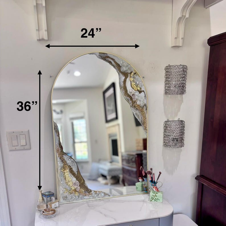 May include: An arched wall mirror with a gold frame, featuring decorative resin accents in white, gray, and gold leaf along the top right and bottom left corners. The mirror measures 24 inches in width and 36 inches in height as indicated.
