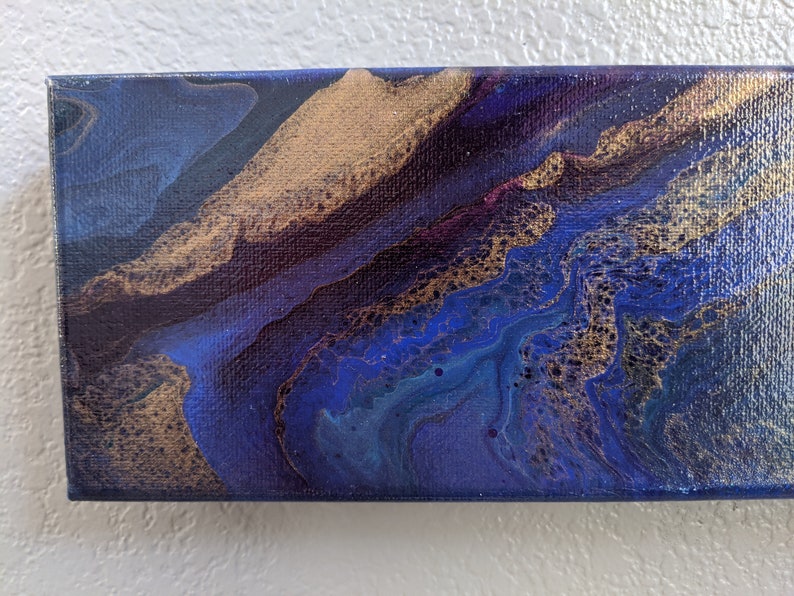 Puede incluir: Pintura abstracta sobre lienzo con remolinos azules, dorados y morados. La pintura tiene una superficie texturizada y un estilo moderno.