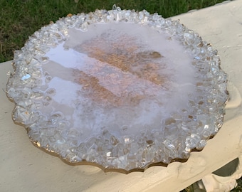 May include: A round, decorative tray with a gold-colored rim. The tray's surface is white with gold flecks and is surrounded by clear, irregularly shaped glass pieces. The tray is sitting on a white surface with green grass in the background.