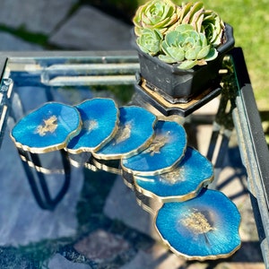 May include: Set of six blue and gold resin coasters with a unique agate design. Each coaster has a gold rim and features gold flecks throughout the blue resin.