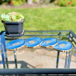 May include: Four blue and gold resin coasters with a wavy edge. The coasters are arranged in a row on a glass table.