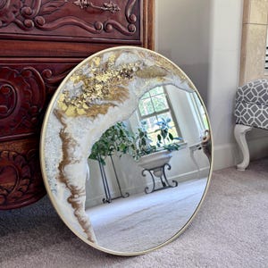 May include: A round wall mirror featuring a gold-toned frame and a decorative resin border. The border displays a swirling pattern of white, cream, and metallic gold pigments, accented with textured gold leaf flakes and shimmering silver glitter.