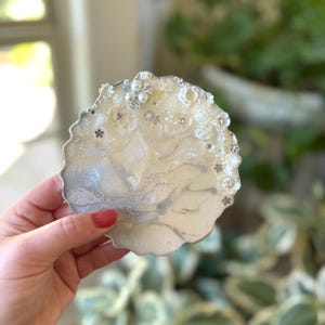 May include: A decorative, round coaster with a silver edge. The coaster has a creamy white and silver marbled design, embellished with small white flowers, pearls, and rhinestones. The coaster is held in a person's hand.