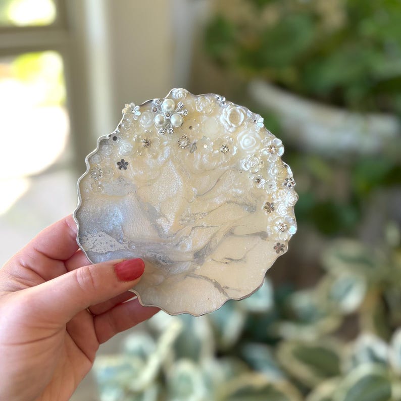 May include: A decorative, round coaster with a silver edge. The coaster has a creamy white and silver marbled design, embellished with small white flowers, pearls, and rhinestones. The coaster is held in a person's hand.