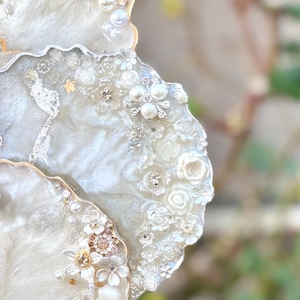 May include: Close-up of a decorative coaster with a pearlescent white resin base. The coaster is adorned with white floral embellishments, faux pearls, and sparkling rhinestones. The edge is trimmed with gold. The design is elegant and suitable for home decor.