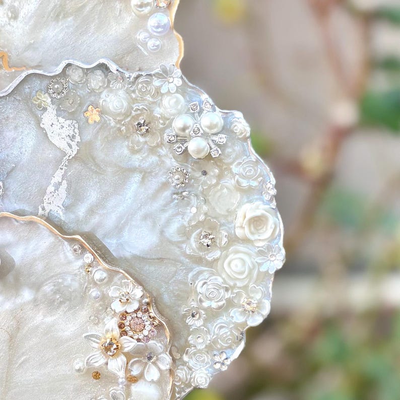 May include: Close-up of a decorative coaster with a pearlescent white resin base. The coaster is adorned with white floral embellishments, faux pearls, and sparkling rhinestones. The edge is trimmed with gold. The design is elegant and suitable for home decor.