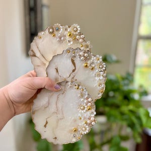 May include: Two decorative resin coasters with a white and gold marbled design. The coasters are embellished with small white flowers, pearls, and gold accents. The edges have a natural, organic shape.