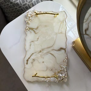 May include: A rectangular decorative tray with a white and gold marbled design. The tray has a clear, textured edge with pearl and floral accents. Gold branch-shaped handles are attached to the tray.
