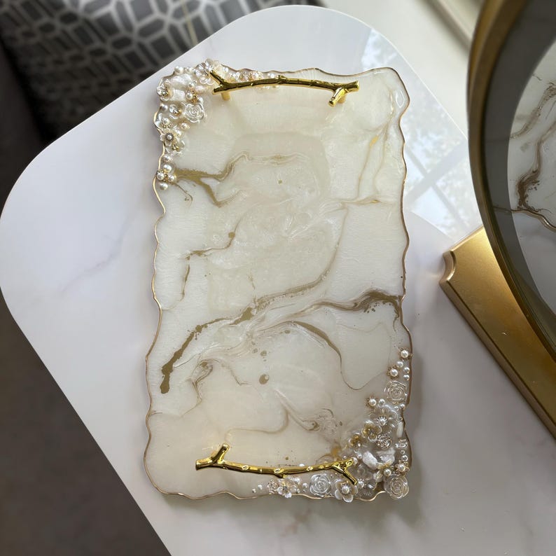 May include: A rectangular decorative tray with a white and gold marbled design. The tray has a clear, textured edge with pearl and floral accents. Gold branch-shaped handles are attached to the tray.
