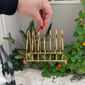 Puede incluir: Portaplatos de metal dorado con seis ranuras. El portaplatos tiene una base redonda y est&aacute; hecho de alambre fino.