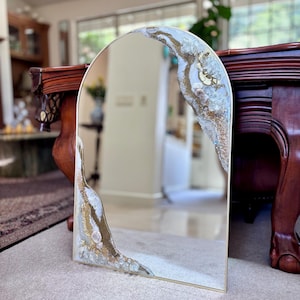 May include: An arched mirror featuring a gold-toned frame and decorative accents along the edges. The design incorporates textured white and clear crystalline elements mixed with shimmering gold glitter, creating a geode-inspired aesthetic.