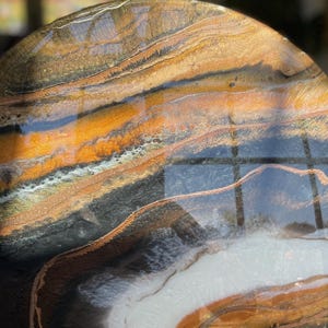 Op de afbeelding: Een rond kunstwerk van hars met een draaiende patroon van zwart, wit, goud en oranje. De hars is helder en glanzend, en de kleuren zijn levendig.