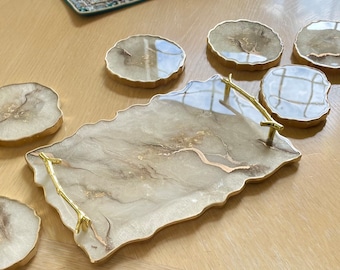 May include: A set of six gold-rimmed, round, agate-style coasters and a matching serving tray with a gold-toned, twig-style handle. The tray and coasters have a white and brown marbled design with gold accents.