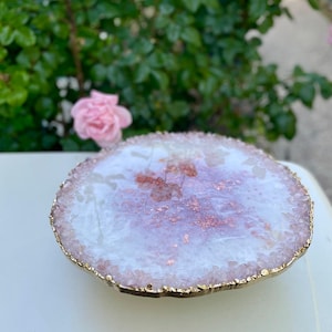 May include: A white agate coaster with a gold rim and pink and copper flecks. The coaster is round and has a natural, irregular edge.