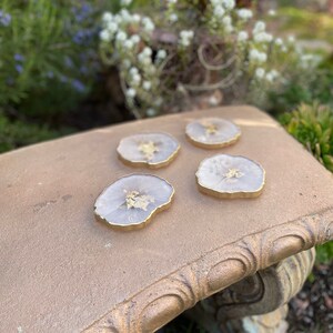 May include: Four white agate coasters with gold leaf accents and a gold rim. The coasters are on a gold-painted surface.