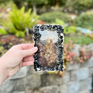 May include: A rectangular resin coaster with a black and gold design. The coaster is surrounded by black crystals.
