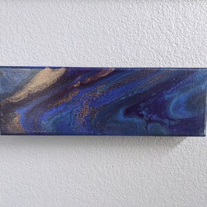 Puede incluir: Pintura abstracta sobre lienzo con colores azul, morado y dorado. La pintura tiene un dise&ntilde;o fluido y arremolinado.