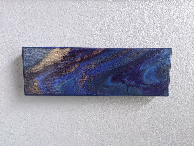 Puede incluir: Pintura abstracta sobre lienzo con colores azul, morado y dorado. La pintura tiene un dise&ntilde;o fluido y arremolinado.