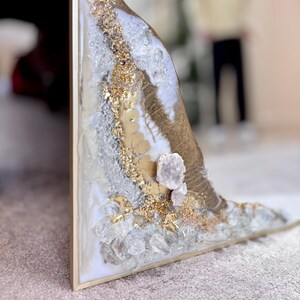 May include: A triangular wall art piece featuring a white resin base with gold leaf accents, embedded clear glass crystals, and a cluster of light purple amethyst geode stones. The artwork is encased in a thin, metallic gold-colored frame.