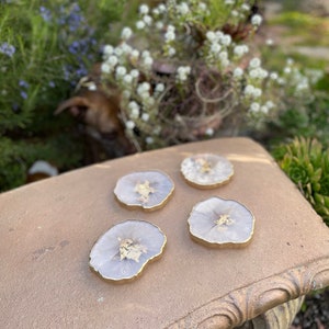 May include: Four round, white agate coasters with gold flecks and a gold rim. The coasters are on a gold-colored surface.