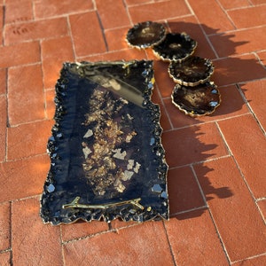 May include: A black and gold resin serving tray with a gold twig handle. The tray has a jagged edge and is decorated with gold leaf. Four matching coasters are also included.