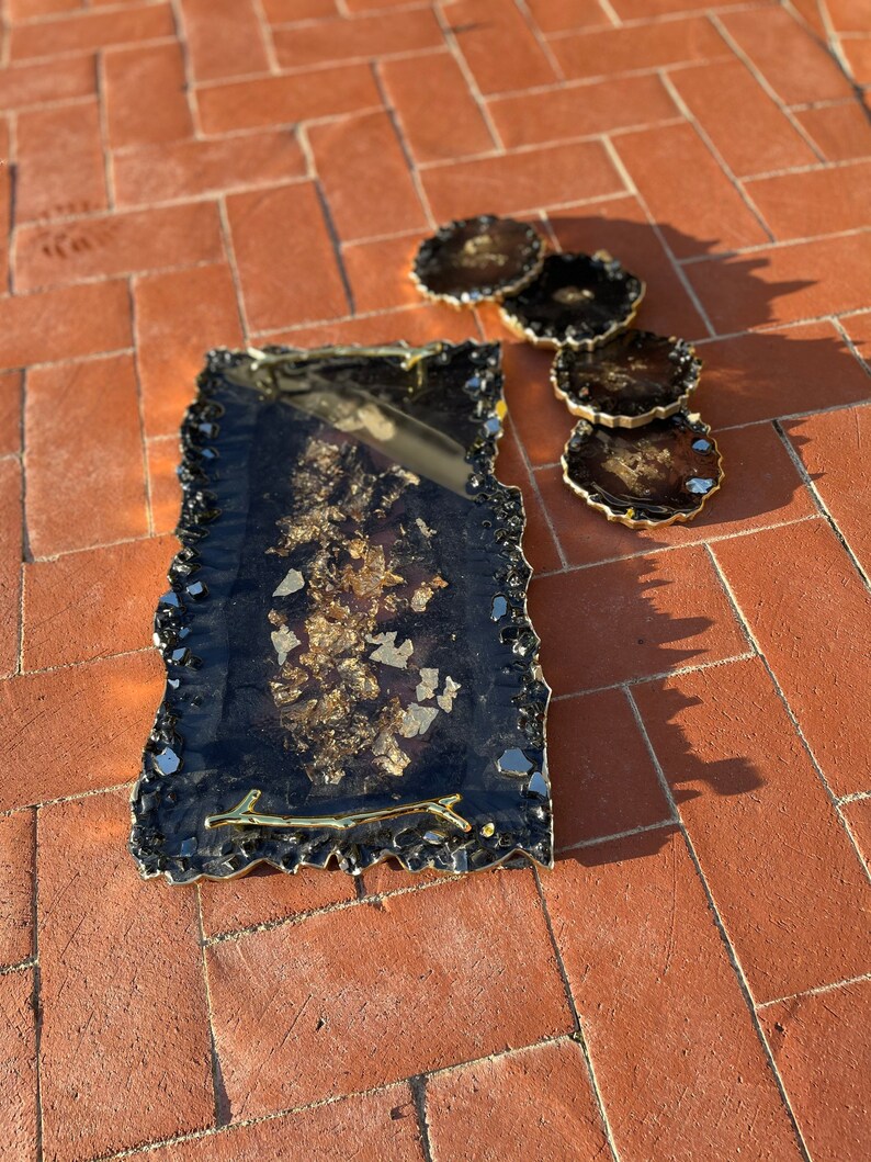May include: A black and gold resin serving tray with a gold twig handle. The tray has a jagged edge and is decorated with gold leaf. Four matching coasters are also included.
