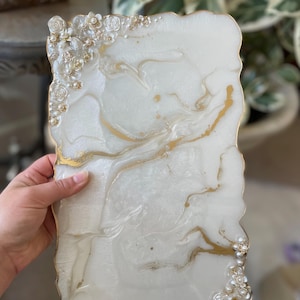 May include: A white resin tray with gold detailing and floral decorations. The tray has a marbled appearance with gold streaks and a gold trim. The edges are irregular, and it features small pearl beads. The tray is held by a hand.