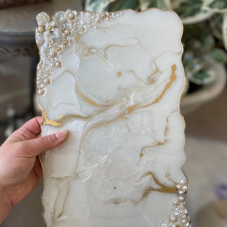 May include: A white resin tray with gold detailing and floral decorations. The tray has a marbled appearance with gold streaks and a gold trim. The edges are irregular, and it features small pearl beads. The tray is held by a hand.