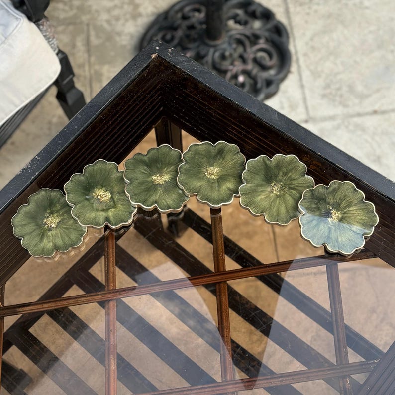 Peut inclure: Six sous-verres en r&eacute;sine verte et dor&eacute;e avec un motif floral. Les sous-verres sont dispos&eacute;s en demi-cercle sur une table en verre.