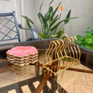 Puede incluir: Un juego de posavasos de color rosa y dorado con forma de flor y un porta servilletas de metal dorado.