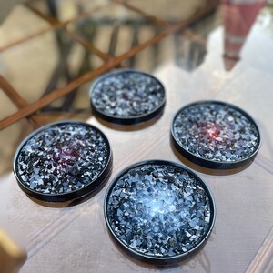 May include: Four black coasters with a glittery, speckled surface. The coasters are round and have a black rim.