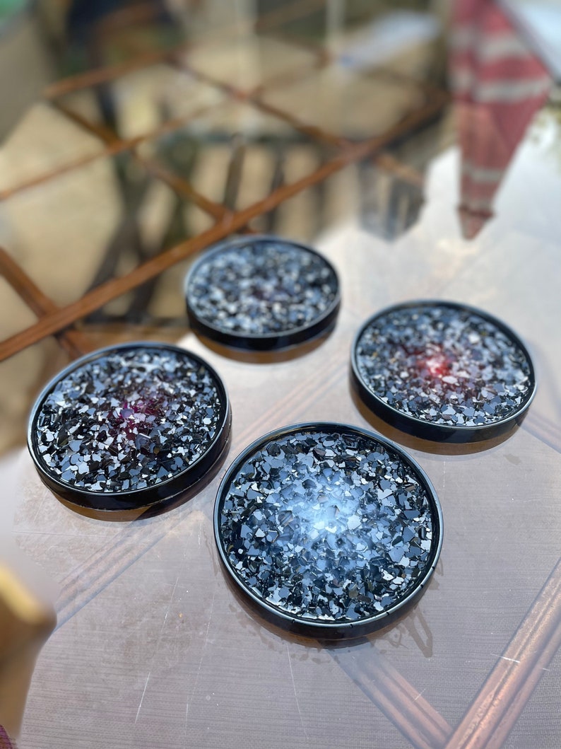 May include: Four black coasters with a glittery, speckled surface. The coasters are round and have a black rim.