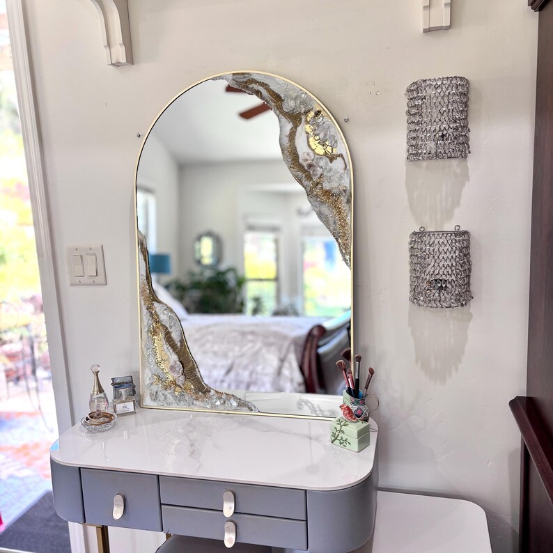 May include: An arched wall mirror with a gold frame featuring decorative resin accents in white, gold, and clear crystal textures along the top right and bottom left edges, positioned above a grey vanity table with a white marble-patterned top.