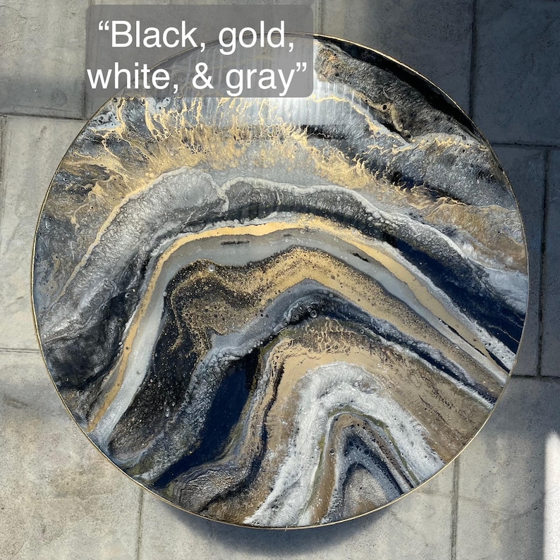 Op de afbeelding: Ronde tafelblad met een abstract ontwerp in zwart, goud, wit en grijs. "Black, gold, white, & gray" staat in witte tekst op de bovenkant van de tafel.